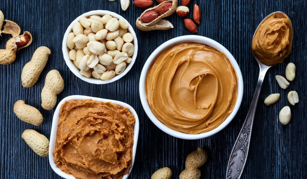 Two bowls of peanut butter and peanuts on dark wooden background from top view