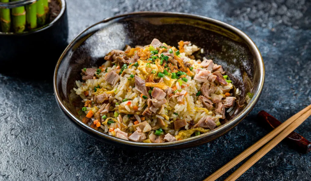 Duck Fried rice in dark bowl on dark stone table with a plant and chopsticks on the side