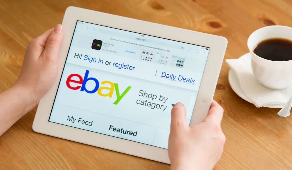 Hand holding an iPad showing Closeup of eBay's website on the table with cup of coffee on the side