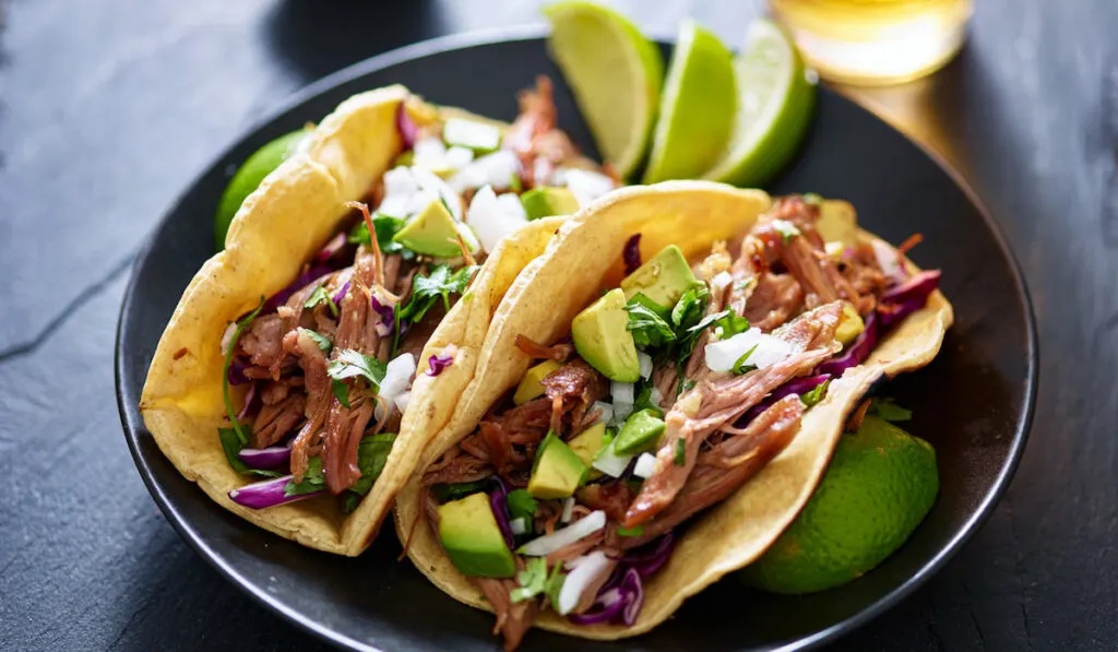 Mexican duck carnita tacos on black plate with lime and beer in background