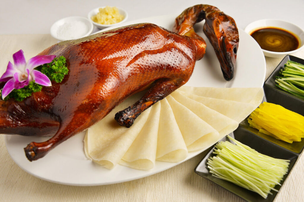 Roasted Whole golden Peking duck plated with wheaten Chinese pancakes, sauce, sliced fresh cucumber, green onions, cilantro and a flower on the table