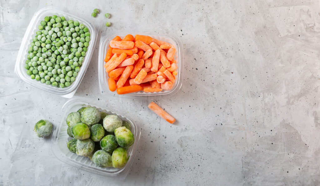 Frozen vegetables such as green peas, brussels sprouts and baby carrots in storage boxes on concrete background