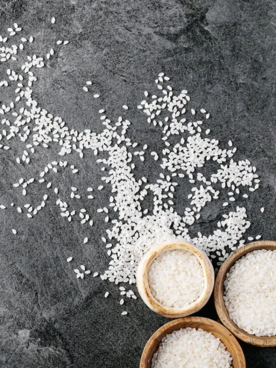 Uncooked rice in wooden and ceramic bowls over dark texture background - ee230526